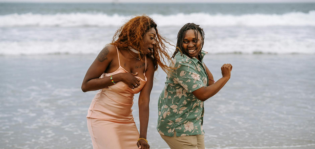 Two Black women laughing and dancing on a beach with ocean waves in the background