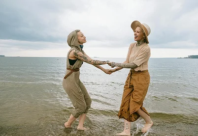 Two happy friends with tattoos holding hands and laughing on a beach to represent healthy emotional attachment.