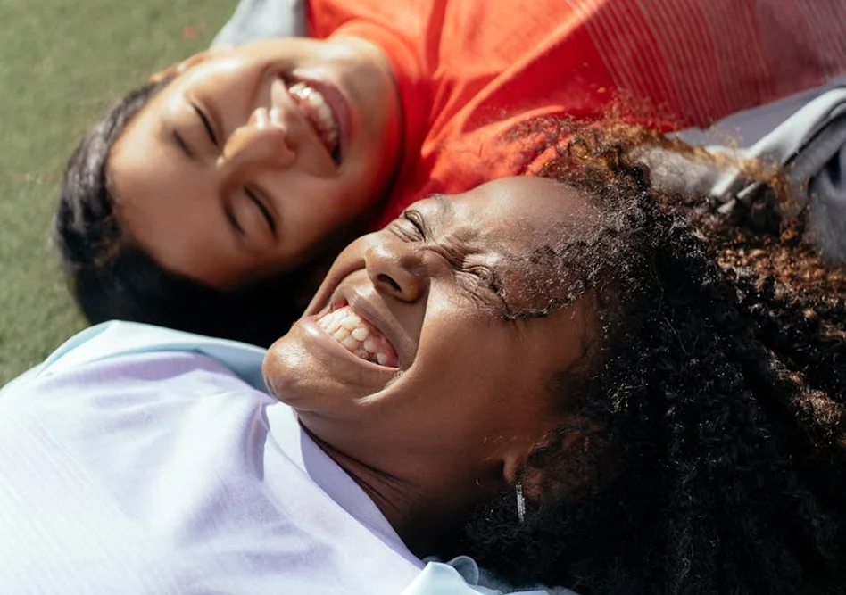 Two young women laughing while lying down on green grass in the sunlight