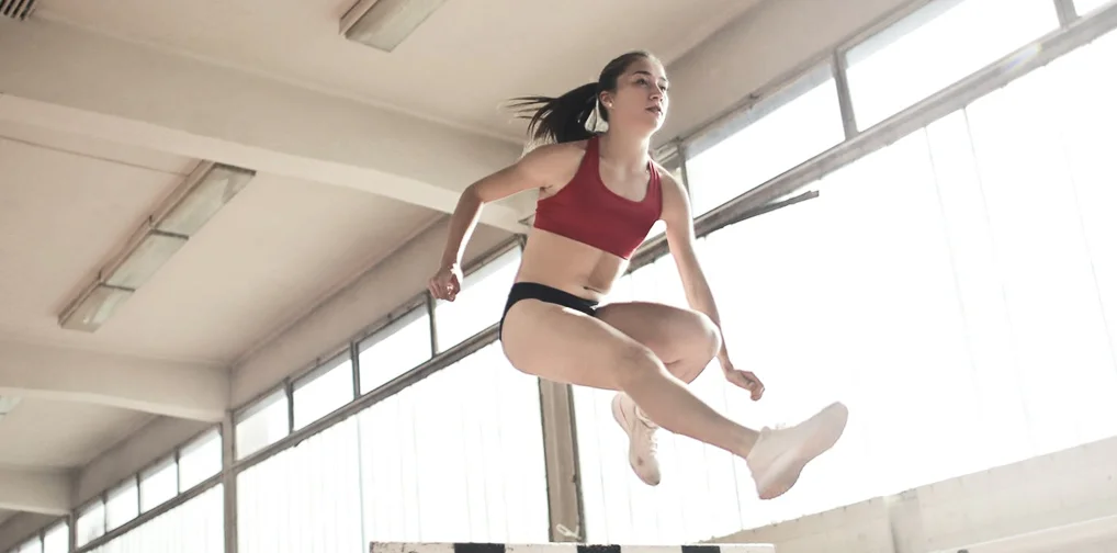 Young female athlete in a red sports bra jumping over a hurdle in an indoor track stadium