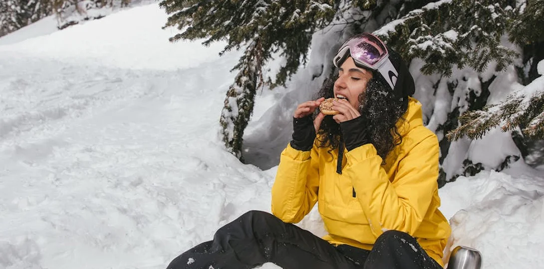 Woman in yellow ski jacket eating a bagel while sitting in the snow under a pine tree