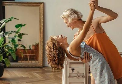 Two women dancing joyfully in a bright, modern living room representing LGBTQ+ affirming therapy and relationship support.