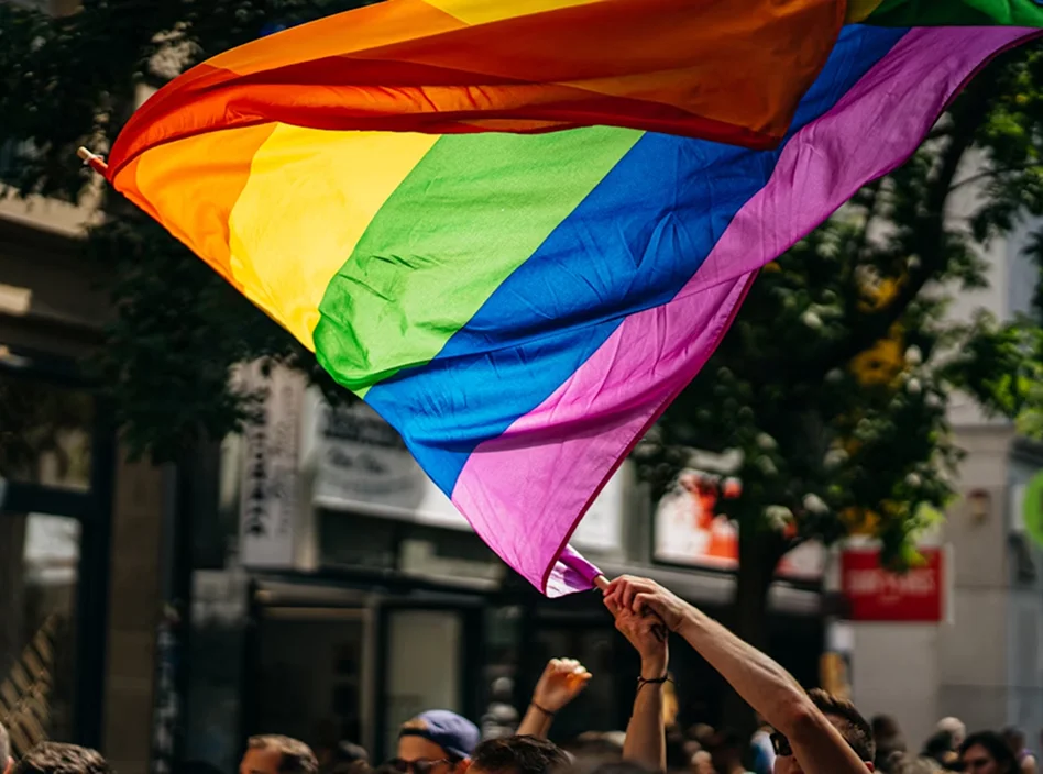 Crowd of people walking down a city street at a Pride parade with rainbow flags draped over their shoulders