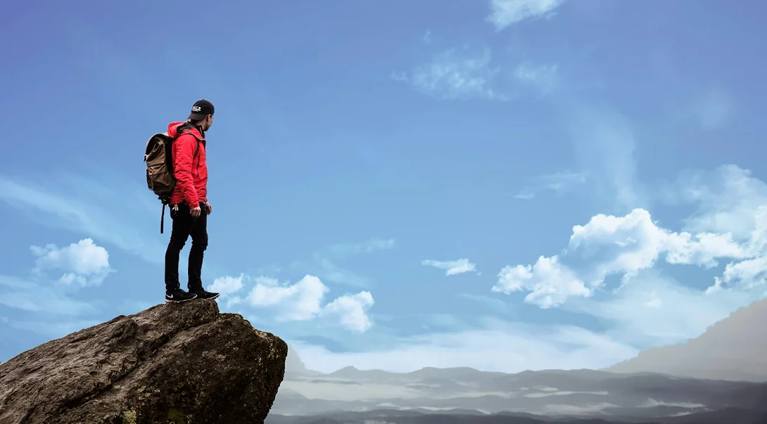 A man in a red jacket with a backpack standing on a rocky mountain peak overlooking a vast valley under a blue sky