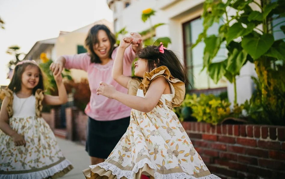 A mother and her two young daughters in floral dresses dancing and laughing together outdoors near sunflowers