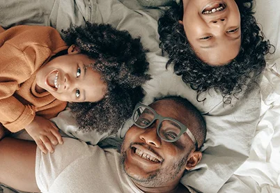 Happy father and two young children laughing together while lying in bed, representing positive parenting and family wellness.