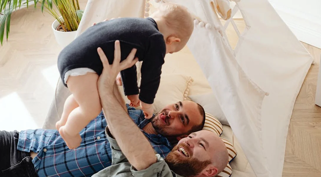Two fathers lying on the floor laughing and playing with their baby inside a play tent