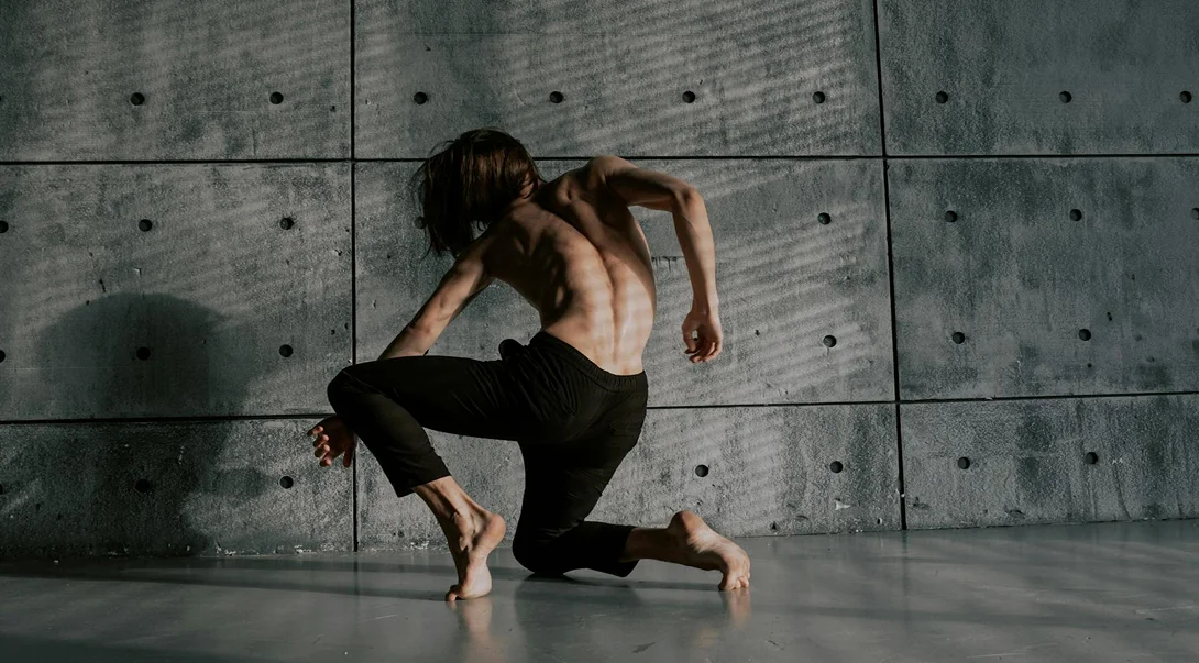 Shirtless person with long hair performing a dramatic, expressive dance pose against a gray concrete wall