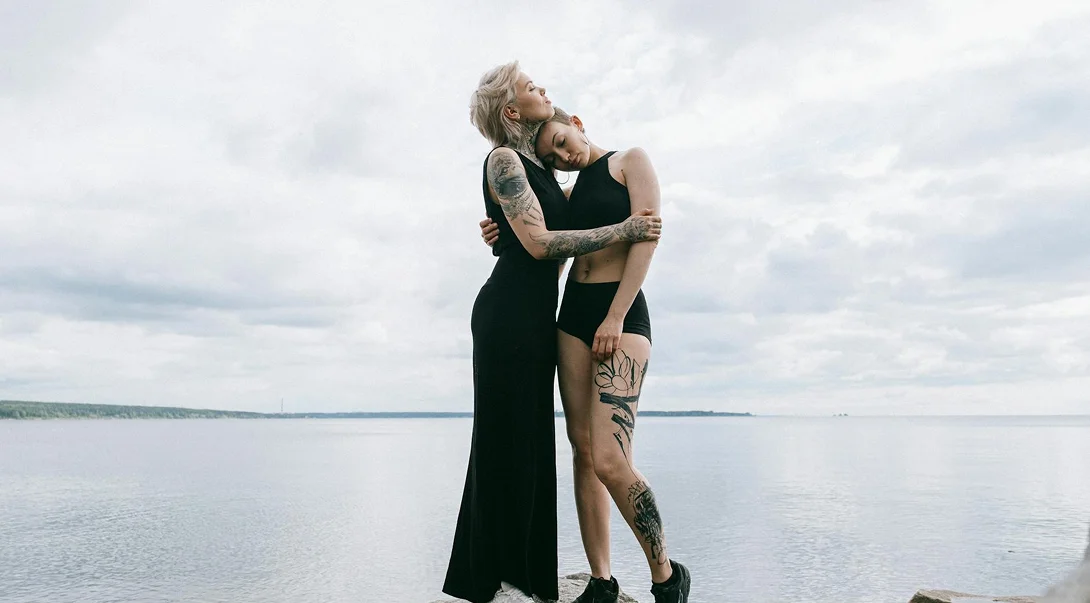 Two women with tattoos embracing supportively on a rocky shore overlooking a calm sea